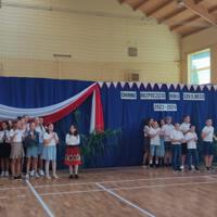 Zdjęcie sali gimnastycznej przedzielonej granatową kotarą na pół. Na środku kotary przymocowany napis GMINNE ROZPOCZĘCIE ROKU SZKOLNEGO 2023-2024. Po lewej stronie opadająca z góry wstęga biało czerwona. Nad napisem i po prawej stronie małe biało czerwone proporczyki przewieszone do drabinek. Dzieci występujące stoją w dwóch licznych grupach. Ubrane są wakacyjnie, chłopcy mają krótkie spodenki a dziewczynki kolorowe spódniczki. Wszystkie mają białe podkoszulki. Jeden chłopiec ubrany jest w strój góralski. Dopełnieniem dekoracji są wysokie trawy i hortensje poukładane w wazonach.