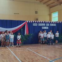Zdjęcie sali gimnastycznej przedzielonej granatową kotarą na pół. Na środku kotary przymocowany napis GMINNE ROZPOCZĘCIE ROKU SZKOLNEGO 2023-2024. Po lewej stronie opadająca z góry wstęga biało czerwona. Nad napisem i po prawej stronie małe biało czerwone proporczyki przewieszone do drabinek. Dzieci występujące stoją w dwóch licznych grupach. Ubrane są wakacyjnie, chłopcy mają krótkie spodenki a dziewczynki kolorowe spódniczki. Wszystkie mają białe podkoszulki. Jeden chłopiec ubrany jest w strój góralski. Dopełnieniem dekoracji są wysokie trawy i hortensje poukładane w wazonach.