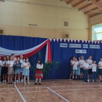 Zdjęcie sali gimnastycznej przedzielonej granatową kotarą na pół. Na środku kotary przymocowany napis GMINNE ROZPOCZĘCIE ROKU SZKOLNEGO 2023-2024. Po lewej stronie opadająca z góry wstęga biało czerwona. Nad napisem i po prawej stronie małe biało czerwone proporczyki przewieszone do drabinek. Dzieci występujące stoją w dwóch licznych grupach. Ubrane są wakacyjnie, chłopcy mają krótkie spodenki a dziewczynki kolorowe spódniczki. Wszystkie mają białe podkoszulki. Jeden chłopiec ubrany jest w strój góralski. Dopełnieniem dekoracji są wysokie trawy i hortensje poukładane w wazonach.