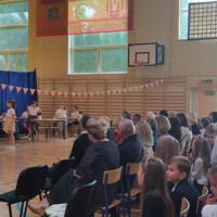 Dzieci recytują wiersze do mikrofonu. Pozostałe siedzą w ławkach, które są ustawione z prawej strony sali. Nad nimi proporczyki.