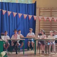 Dzieci recytują wiersze do mikrofonu. Pozostałe siedzą w ławkach, które są ustawione z prawej strony sali. Nad nimi proporczyki.