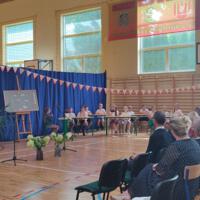 Dzieci recytują wiersze do mikrofonu. Pozostałe siedzą w ławkach, które są ustawione z prawej strony sali. Nad nimi proporczyki.