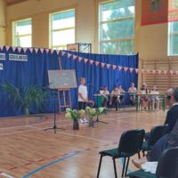 Dzieci recytują wiersze do mikrofonu. Pozostałe siedzą w ławkach, które są ustawione z prawej strony sali. Nad nimi proporczyki.