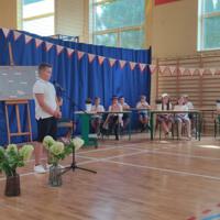 Dzieci recytują wiersze do mikrofonu. Pozostałe siedzą w ławkach, które są ustawione z prawej strony sali. Nad nimi proporczyki.