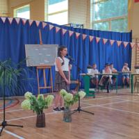 Dzieci recytują wiersze do mikrofonu. Pozostałe siedzą w ławkach, które są ustawione z prawej strony sali. Nad nimi proporczyki.