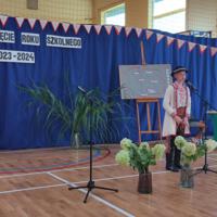 Dzieci recytują wiersze do mikrofonu. Pozostałe siedzą w ławkach, które są ustawione z prawej strony sali. Nad nimi proporczyki.