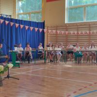 Dzieci recytują wiersze do mikrofonu. Pozostałe siedzą w ławkach, które są ustawione z prawej strony sali. Nad nimi proporczyki.