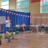 Dzieci recytują wiersze do mikrofonu. Pozostałe siedzą w ławkach, które są ustawione z prawej strony sali. Nad nimi proporczyki.