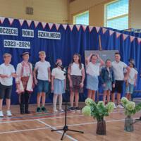 Dzieci recytują wiersze do mikrofonu. Pozostałe siedzą w ławkach, które są ustawione z prawej strony sali. Nad nimi proporczyki.