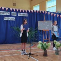 Dzieci recytują wiersze do mikrofonu. Pozostałe siedzą w ławkach, które są ustawione z prawej strony sali. Nad nimi proporczyki.