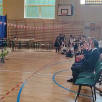 Dzieci recytują wiersze do mikrofonu. Pozostałe siedzą w ławkach, które są ustawione z prawej strony sali. Nad nimi proporczyki.