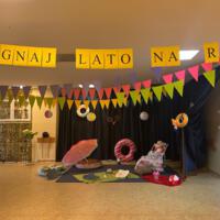 Zdjęcie dekoracji korytarza utrzymane w stylu wakacyjnym. U góry napis na żółtych kartkach "Żegnaj lato na rok!". Na ziemi oraz na zasłonie akcesoria kojarzone z latem: leżak, koce, koła do pływania.