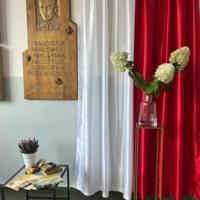 Zdjęcie dekoracji korytarza obok patrona szkoły. Dołożony stoliczek z kałamarzem, starymi gazetami i wrzosem w doniczce. Obok obok na tle barw narodowych hortensje we flakonie.