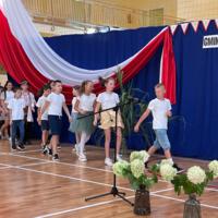 Zdjęcie sali gimnastycznej przedzielonej granatową kotarą na pół. Na środku kotary przymocowany napis GMINNE ROZPOCZĘCIE ROKU SZKOLNEGO 2023-2024. Po lewej stronie opadająca z góry wstęga biało czerwona. Nad napisem i po prawej stronie małe biało czerwone proporczyki przewieszone do drabinek. Dzieci występujące stoją w dwóch licznych grupach. Ubrane są wakacyjnie, chłopcy mają krótkie spodenki a dziewczynki kolorowe spódniczki. Wszystkie mają białe podkoszulki. Jeden chłopiec ubrany jest w strój góralski. Dopełnieniem dekoracji są wysokie trawy i hortensje poukładane w wazonach.