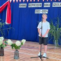 Dzieci recytują wiersze do mikrofonu. Pozostałe siedzą w ławkach, które są ustawione z prawej strony sali. Nad nimi proporczyki.