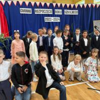 Dzieci recytują wiersze do mikrofonu. Pozostałe siedzą w ławkach, które są ustawione z prawej strony sali. Nad nimi proporczyki.