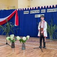 Dzieci recytują wiersze do mikrofonu. Pozostałe siedzą w ławkach, które są ustawione z prawej strony sali. Nad nimi proporczyki.