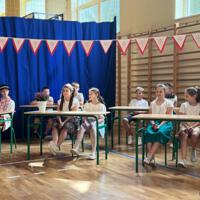 Dzieci recytują wiersze do mikrofonu. Pozostałe siedzą w ławkach, które są ustawione z prawej strony sali. Nad nimi proporczyki.