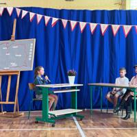 Dzieci recytują wiersze do mikrofonu. Pozostałe siedzą w ławkach, które są ustawione z prawej strony sali. Nad nimi proporczyki.