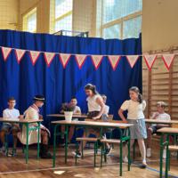 Dzieci recytują wiersze do mikrofonu. Pozostałe siedzą w ławkach, które są ustawione z prawej strony sali. Nad nimi proporczyki.