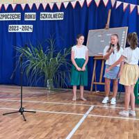 Dzieci recytują wiersze do mikrofonu. Pozostałe siedzą w ławkach, które są ustawione z prawej strony sali. Nad nimi proporczyki.