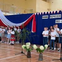 Zdjęcie sali gimnastycznej przedzielonej granatową kotarą na pół. Na środku kotary przymocowany napis GMINNE ROZPOCZĘCIE ROKU SZKOLNEGO 2023-2024. Po lewej stronie opadająca z góry wstęga biało czerwona. Nad napisem i po prawej stronie małe biało czerwone proporczyki przewieszone do drabinek. Dzieci występujące stoją w dwóch licznych grupach. Ubrane są wakacyjnie, chłopcy mają krótkie spodenki a dziewczynki kolorowe spódniczki. Wszystkie mają białe podkoszulki. Jeden chłopiec ubrany jest w strój góralski. Dopełnieniem dekoracji są wysokie trawy i hortensje poukładane w wazonach.