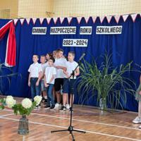 Zdjęcie sali gimnastycznej przedzielonej granatową kotarą na pół. Na środku kotary przymocowany napis GMINNE ROZPOCZĘCIE ROKU SZKOLNEGO 2023-2024. Po lewej stronie opadająca z góry wstęga biało czerwona. Nad napisem i po prawej stronie małe biało czerwone proporczyki przewieszone do drabinek. Dzieci występujące stoją w dwóch licznych grupach. Ubrane są wakacyjnie, chłopcy mają krótkie spodenki a dziewczynki kolorowe spódniczki. Wszystkie mają białe podkoszulki. Jeden chłopiec ubrany jest w strój góralski. Dopełnieniem dekoracji są wysokie trawy i hortensje poukładane w wazonach.