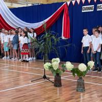 Zdjęcie sali gimnastycznej przedzielonej granatową kotarą na pół. Na środku kotary przymocowany napis GMINNE ROZPOCZĘCIE ROKU SZKOLNEGO 2023-2024. Po lewej stronie opadająca z góry wstęga biało czerwona. Nad napisem i po prawej stronie małe biało czerwone proporczyki przewieszone do drabinek. Dzieci występujące stoją w dwóch licznych grupach. Ubrane są wakacyjnie, chłopcy mają krótkie spodenki a dziewczynki kolorowe spódniczki. Wszystkie mają białe podkoszulki. Jeden chłopiec ubrany jest w strój góralski. Dopełnieniem dekoracji są wysokie trawy i hortensje poukładane w wazonach.