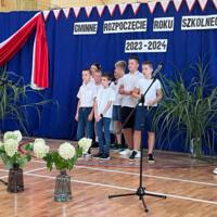 Zdjęcie sali gimnastycznej przedzielonej granatową kotarą na pół. Na środku kotary przymocowany napis GMINNE ROZPOCZĘCIE ROKU SZKOLNEGO 2023-2024. Po lewej stronie opadająca z góry wstęga biało czerwona. Nad napisem i po prawej stronie małe biało czerwone proporczyki przewieszone do drabinek. Dzieci występujące stoją w dwóch licznych grupach. Ubrane są wakacyjnie, chłopcy mają krótkie spodenki a dziewczynki kolorowe spódniczki. Wszystkie mają białe podkoszulki. Jeden chłopiec ubrany jest w strój góralski. Dopełnieniem dekoracji są wysokie trawy i hortensje poukładane w wazonach.