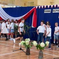 Zdjęcie sali gimnastycznej przedzielonej granatową kotarą na pół. Na środku kotary przymocowany napis GMINNE ROZPOCZĘCIE ROKU SZKOLNEGO 2023-2024. Po lewej stronie opadająca z góry wstęga biało czerwona. Nad napisem i po prawej stronie małe biało czerwone proporczyki przewieszone do drabinek. Dzieci występujące stoją w dwóch licznych grupach. Ubrane są wakacyjnie, chłopcy mają krótkie spodenki a dziewczynki kolorowe spódniczki. Wszystkie mają białe podkoszulki. Jeden chłopiec ubrany jest w strój góralski. Dopełnieniem dekoracji są wysokie trawy i hortensje poukładane w wazonach.