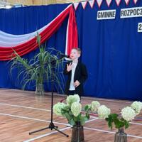 Dzieci recytują wiersze do mikrofonu. Pozostałe siedzą w ławkach, które są ustawione z prawej strony sali. Nad nimi proporczyki.