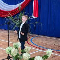 Dzieci recytują wiersze do mikrofonu. Pozostałe siedzą w ławkach, które są ustawione z prawej strony sali. Nad nimi proporczyki.