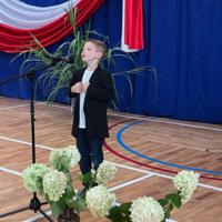 Dzieci recytują wiersze do mikrofonu. Pozostałe siedzą w ławkach, które są ustawione z prawej strony sali. Nad nimi proporczyki.