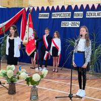 Na zdjęciu dwie uczennice prowadzące uroczystość. Za nimi trójka dzieci niosąca sztandar. na tle dekoracji. 