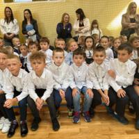 Na zdjęciach uczniowie szkoły, którzy oglądali akademię. Młodsze dzieci siedzą na ławeczkach. Starsze na krzesłach, najstarsi stoją z tyłu. Wszyscy są odświętnie ubrani.