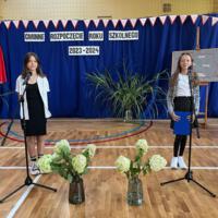 Dzieci recytują wiersze do mikrofonu. Pozostałe siedzą w ławkach, które są ustawione z prawej strony sali. Nad nimi proporczyki.