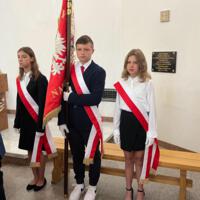 Na zdjęciu dwie uczennice oraz pomiędzy nimi uczeń trzymający sztandar. Wszyscy ubrani galowo, na dłonie mają założone rękawiczki.