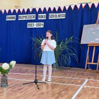 Dzieci recytują wiersze do mikrofonu. Pozostałe siedzą w ławkach, które są ustawione z prawej strony sali. Nad nimi proporczyki.