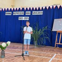 Dzieci recytują wiersze do mikrofonu. Pozostałe siedzą w ławkach, które są ustawione z prawej strony sali. Nad nimi proporczyki.