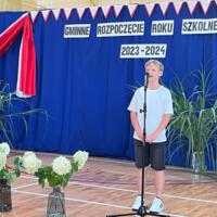 Dzieci recytują wiersze do mikrofonu. Pozostałe siedzą w ławkach, które są ustawione z prawej strony sali. Nad nimi proporczyki.