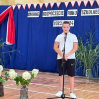 Dzieci recytują wiersze do mikrofonu. Pozostałe siedzą w ławkach, które są ustawione z prawej strony sali. Nad nimi proporczyki.