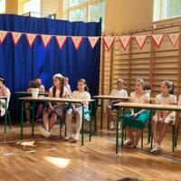 Dzieci recytują wiersze do mikrofonu. Pozostałe siedzą w ławkach, które są ustawione z prawej strony sali. Nad nimi proporczyki.