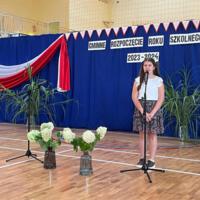 Dzieci recytują wiersze do mikrofonu. Pozostałe siedzą w ławkach, które są ustawione z prawej strony sali. Nad nimi proporczyki.