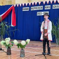 Dzieci recytują wiersze do mikrofonu. Pozostałe siedzą w ławkach, które są ustawione z prawej strony sali. Nad nimi proporczyki.