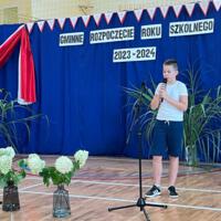 Dzieci recytują wiersze do mikrofonu. Pozostałe siedzą w ławkach, które są ustawione z prawej strony sali. Nad nimi proporczyki.