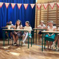 Dzieci recytują wiersze do mikrofonu. Pozostałe siedzą w ławkach, które są ustawione z prawej strony sali. Nad nimi proporczyki.