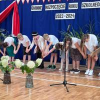 Dzieci recytują wiersze do mikrofonu. Pozostałe siedzą w ławkach, które są ustawione z prawej strony sali. Nad nimi proporczyki.