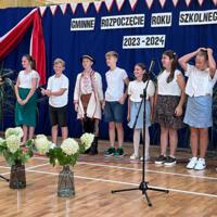 Dzieci recytują wiersze do mikrofonu. Pozostałe siedzą w ławkach, które są ustawione z prawej strony sali. Nad nimi proporczyki.