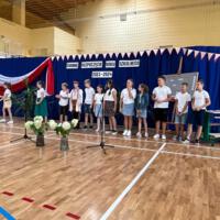 Zdjęcie sali gimnastycznej przedzielonej granatową kotarą na pół. Na środku kotary przymocowany napis GMINNE ROZPOCZĘCIE ROKU SZKOLNEGO 2023-2024. Po lewej stronie opadająca z góry wstęga biało czerwona. Nad napisem i po prawej stronie małe biało czerwone proporczyki przewieszone do drabinek. Dzieci występujące stoją w dwóch licznych grupach. Ubrane są wakacyjnie, chłopcy mają krótkie spodenki a dziewczynki kolorowe spódniczki. Wszystkie mają białe podkoszulki. Jeden chłopiec ubrany jest w strój góralski. Dopełnieniem dekoracji są wysokie trawy i hortensje poukładane w wazonach.