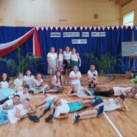 Uczniowie klasy. która brała udział w akademii zapozowali do wspólnego zdjęcia na tle dekoracji. Cztery dziewczynki stoją w centralnej części zdjęcia i trzymają się za ramiona. Pozostałe dzieci siedzą po turecku lub leżą na podłodze. 